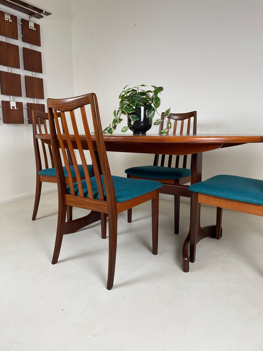 A Teak Gplan Table on a grey floor and in a white walled room, with a midcentury brown wall hanging to the left. The table is surrounded by 4 matching chairs and the table top has a potted plant on top of it.