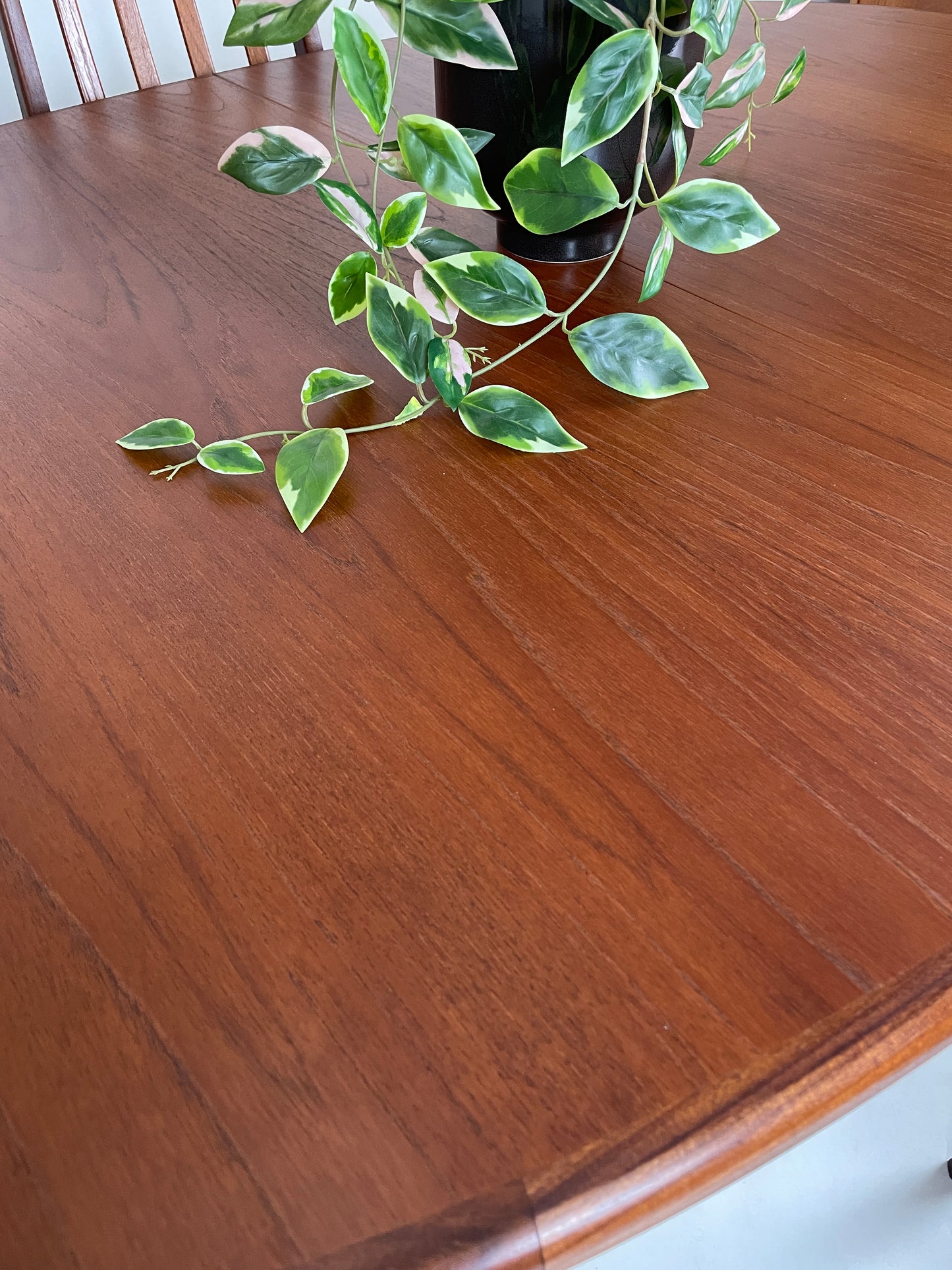 the table top is a beautiful teak colour with grains of wood and has been finished to a high standard with no visible marks or damage. Th e table top has a potted plant in a black vase on top of it and you can see just one matching chair at the other side of the table.