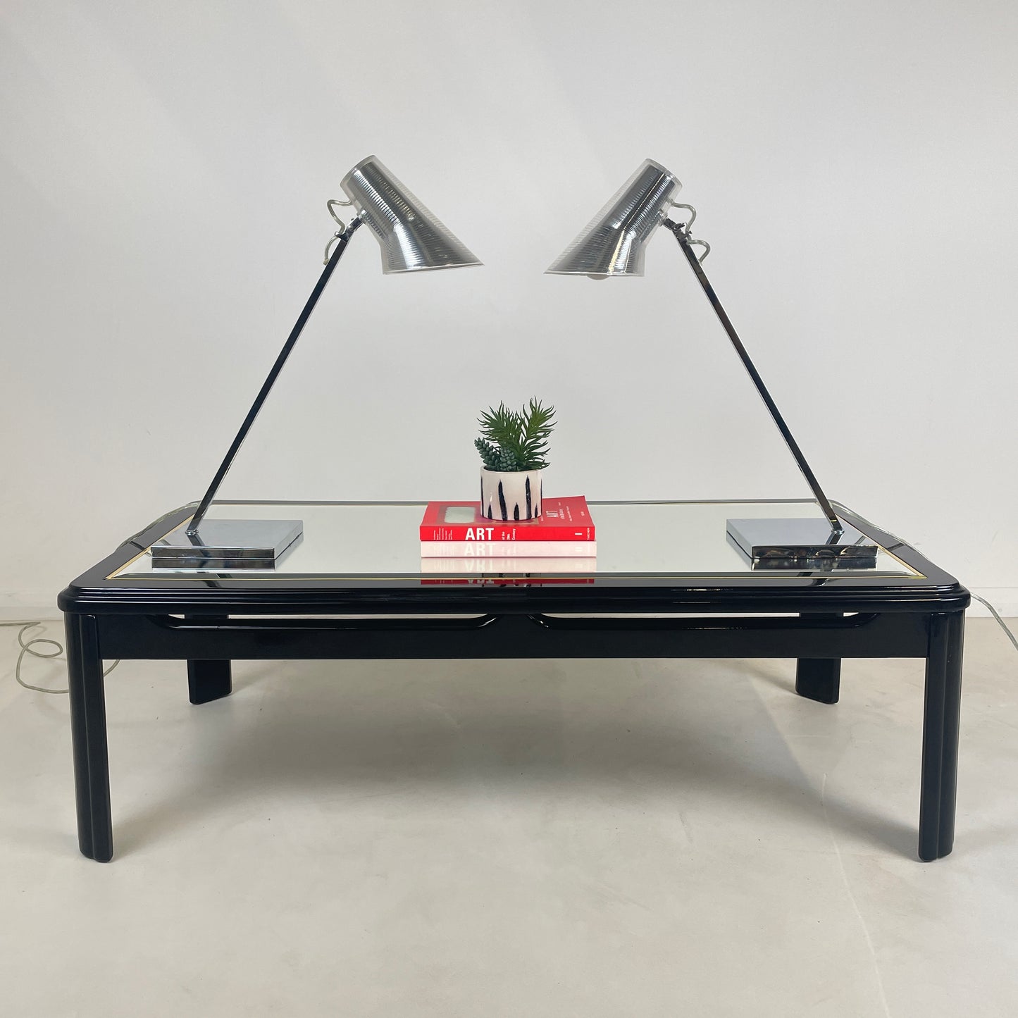 the pair of 2 matching Silver Desk lamps of a vintage glass top coffee table, the lamps are facing each other to they're identical and have two art books in the middle of the lamps with a potted plant above the books.