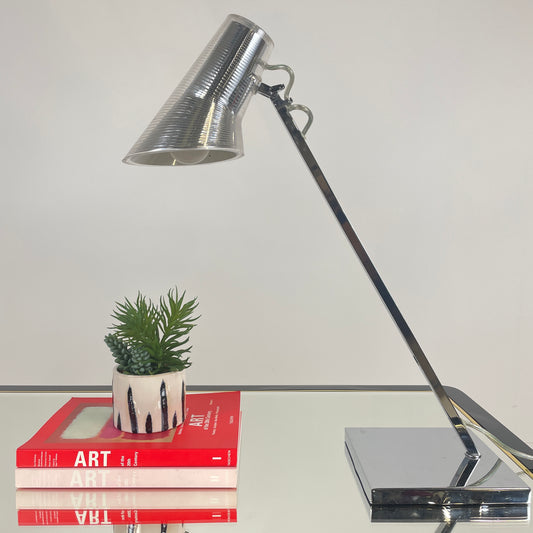 one of the lamps is pictured closer to show its detail, it has a silver top with a clear covering that is corrugated and clear wiring, the bottom of the lamp stand is square and it has a long thin silver neck. It is shown above two art books and a small potted plant.