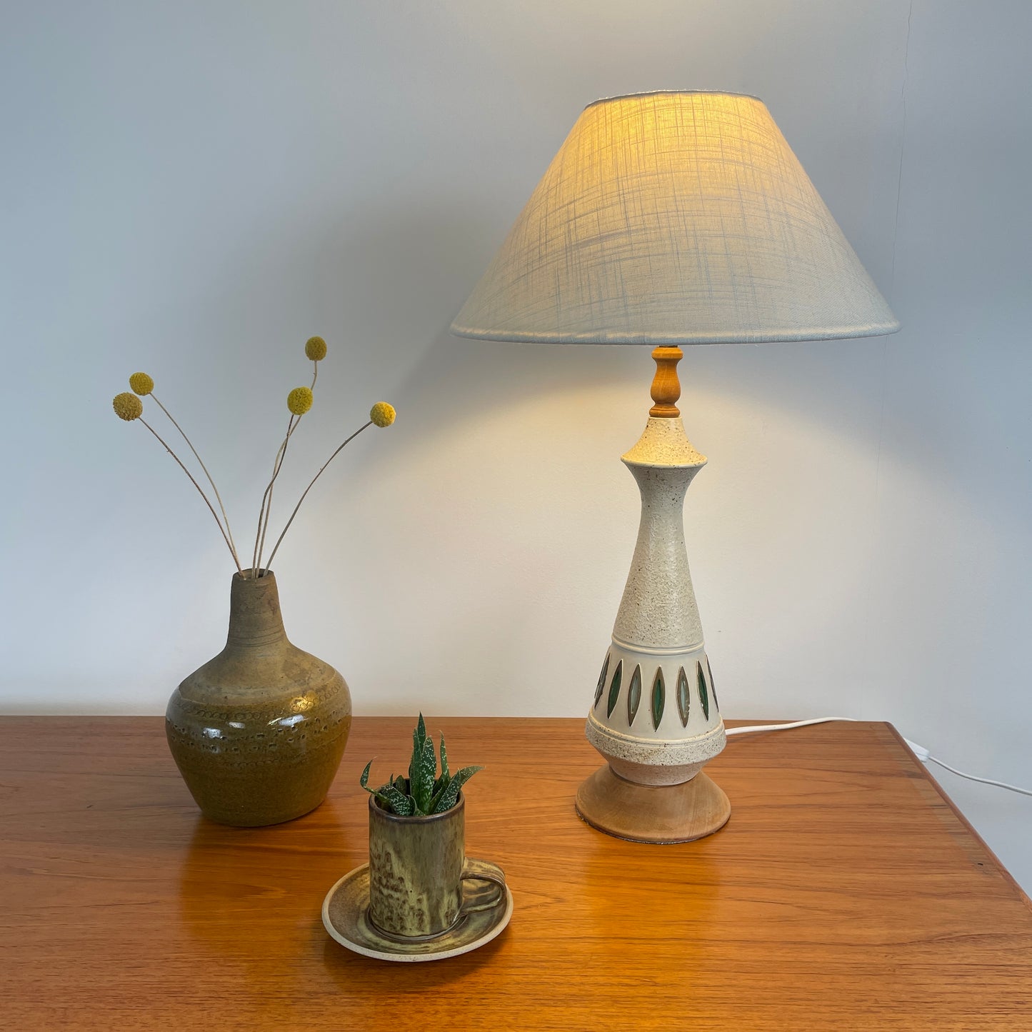 A Midcentury Table Lamp made from wood & pottery with a neutral lampshade, on a teak unit against a white wall. The teak unit also has a retro case with dried flowers in it and a cute retro cup & saucer with a succulent inside