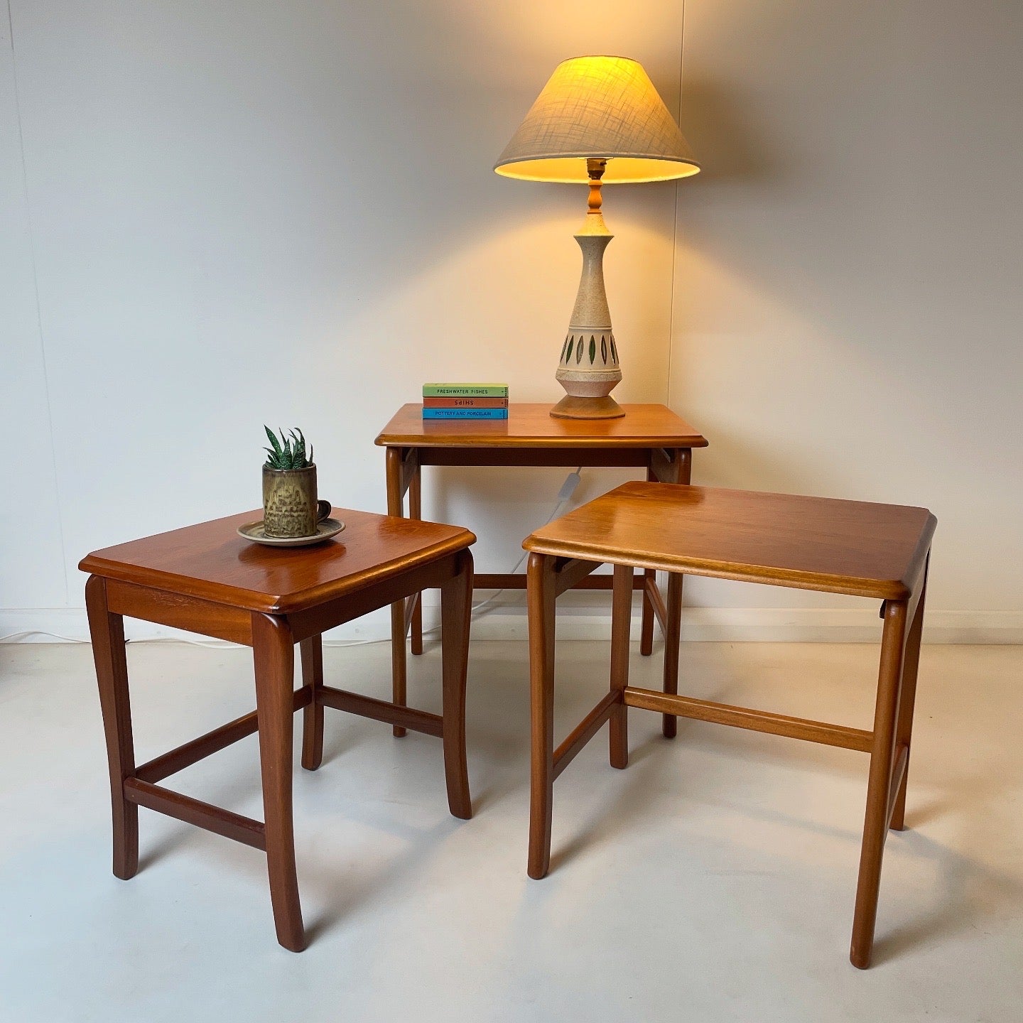 the wood & pottery lamp is on top of a midcentury table that is part of a set of three nest of tables that are also pictured. These are all on a solid concrete floor against a white wall.