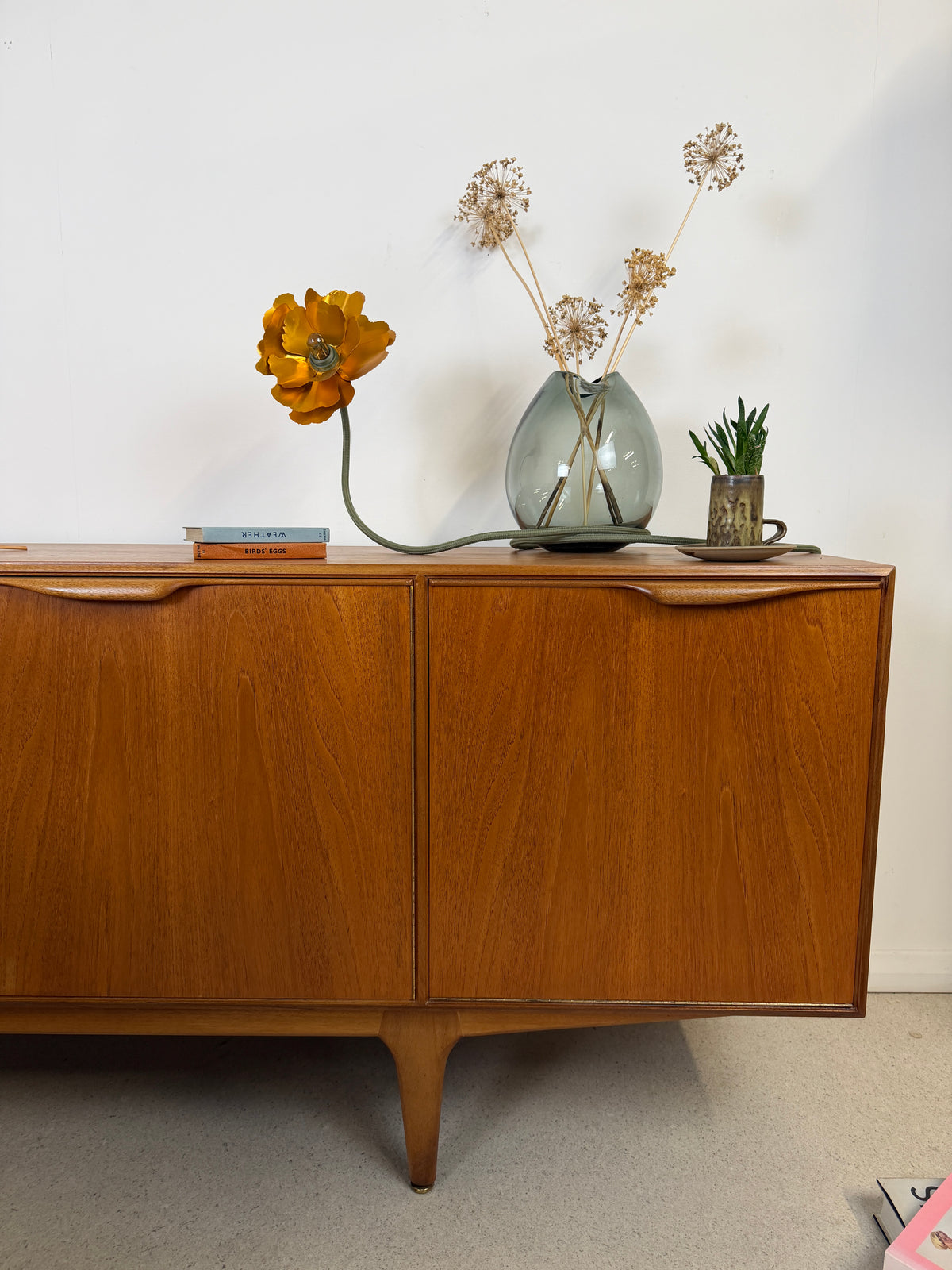 The habitat flower lamp is sat on a large vintage sideboard with a vase of flowers, some books,  a coffee cup that has a plant displayed in it