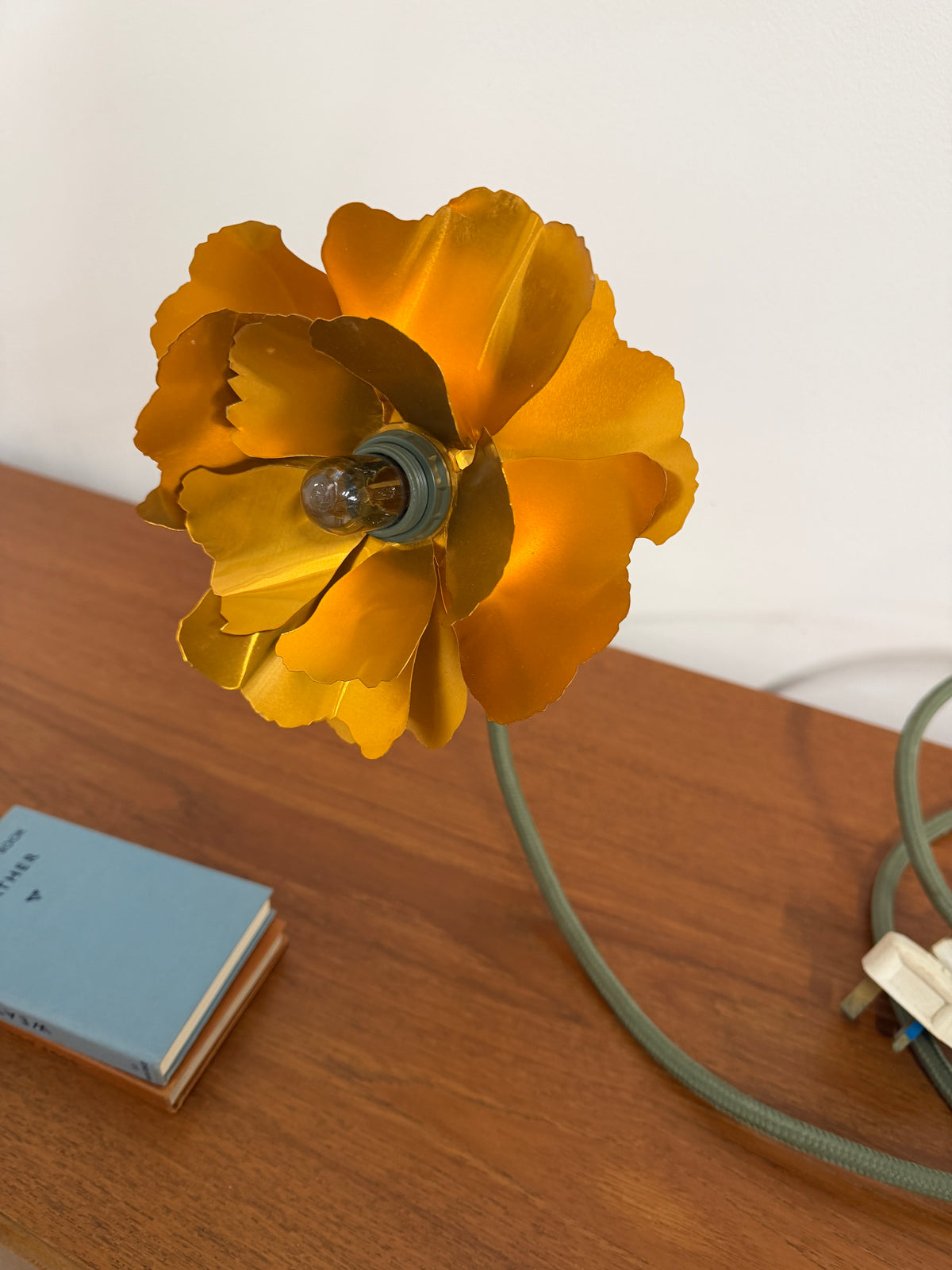 A vintage-style flower-shaped table lamp with bright yellow-orange petals and a green cord, shown resting on a wooden tabletop next to a small blue book.