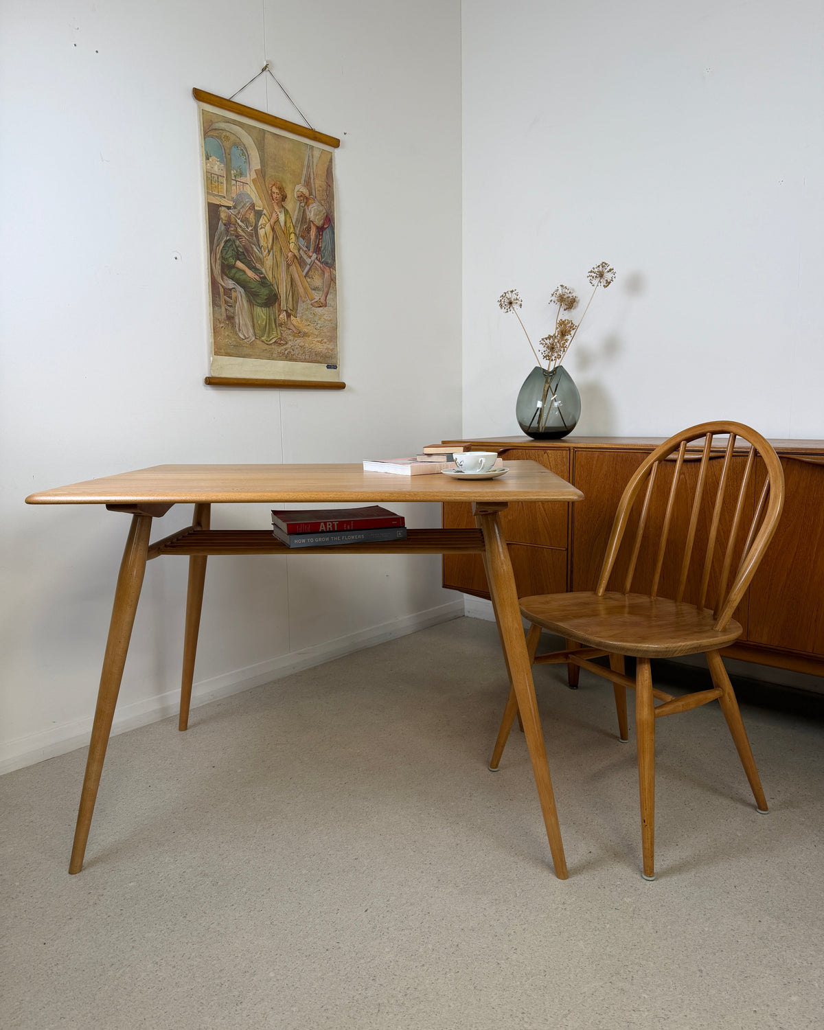 A mid-century modern light wood dining table  with a matching wooden spindle-back chair, set in a minimalist room with a white wall, hanging tapestry, and concrete style floor