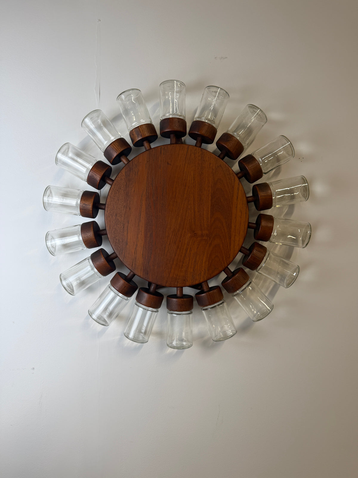 A circular wall-mounted spice  rack with a round medium-brown wooden centre and 18 clear glass jars with wooden lids displayed on a white wall