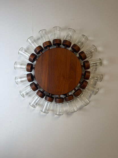 A circular wall-mounted spice  rack with a round medium-brown wooden centre and 18 clear glass jars with wooden lids displayed on a white wall