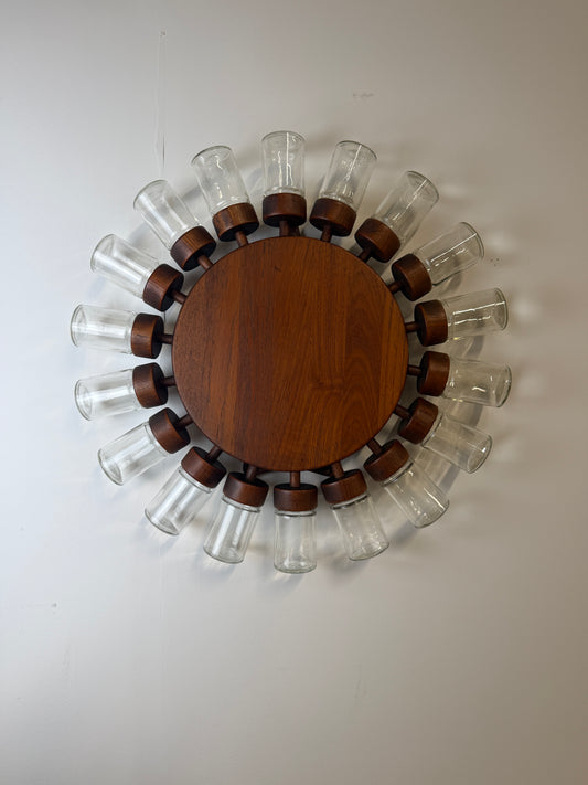 A circular wall-mounted spice  rack with a round medium-brown wooden centre and 18 clear glass jars with wooden lids displayed on a white wall