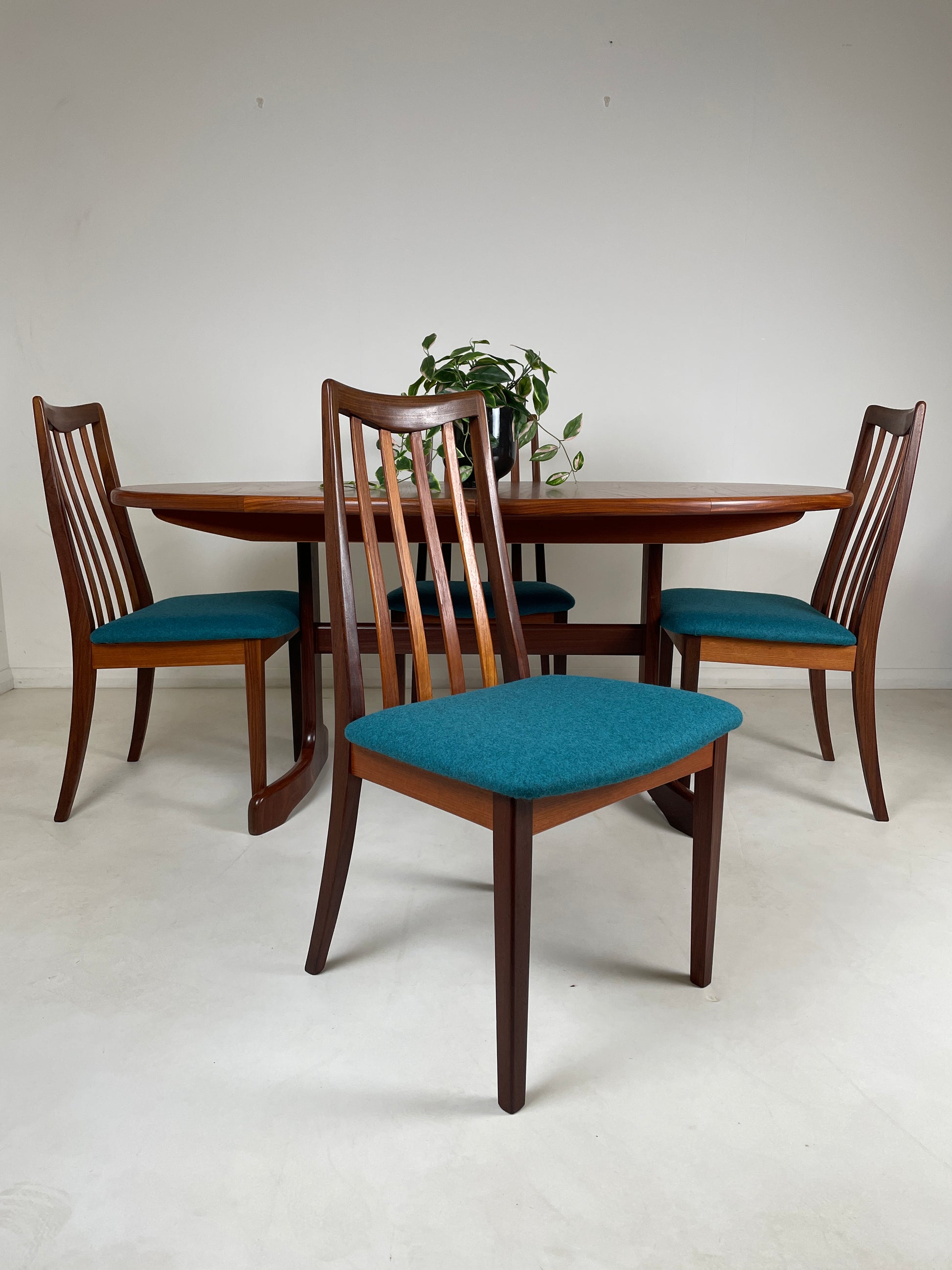 the table is in a white room with grey floors, it has 4 chairs tucked under at each end and side of the table. the closest chair to the foreground is pulled out and faces the camera, you can see the upholstered teal cushion on the chair itself.
