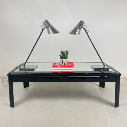 the pair of 2 matching Silver Desk lamps of a vintage glass top coffee table, the lamps are facing each other to they're identical and have two art books in the middle of the lamps with a potted plant above the books. 