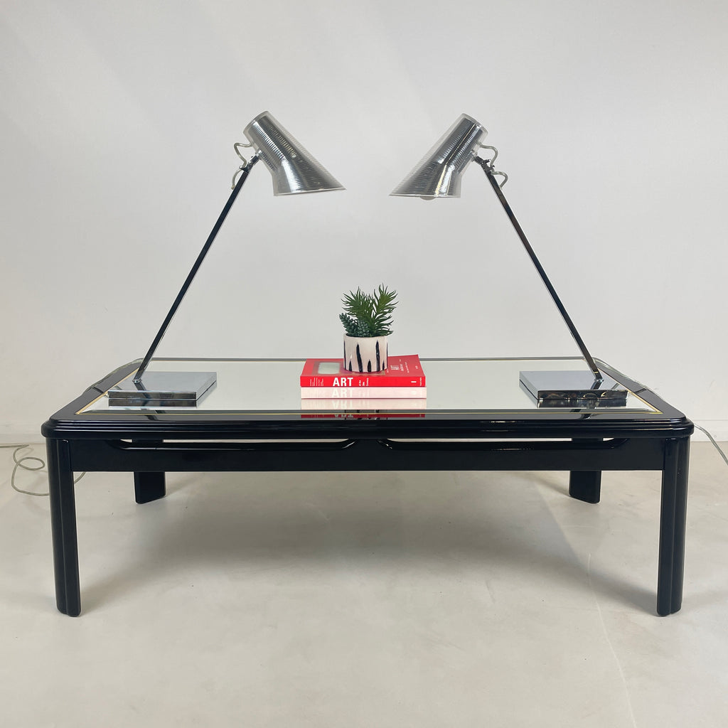 the pair of 2 matching Silver Desk lamps of a vintage glass top coffee table, the lamps are facing each other to they're identical and have two art books in the middle of the lamps with a potted plant above the books. 