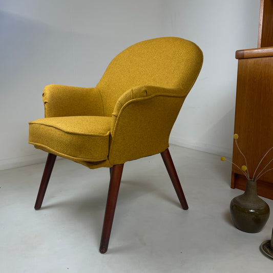 the chair has been upholstered with a mustard colour strong fabric, with teak wooden legs, there is retro vase beside it with dried yellow flowers in and the background has a teak Gplan unit. The room is white which showcases the stunning vibrant colour of the chair itself