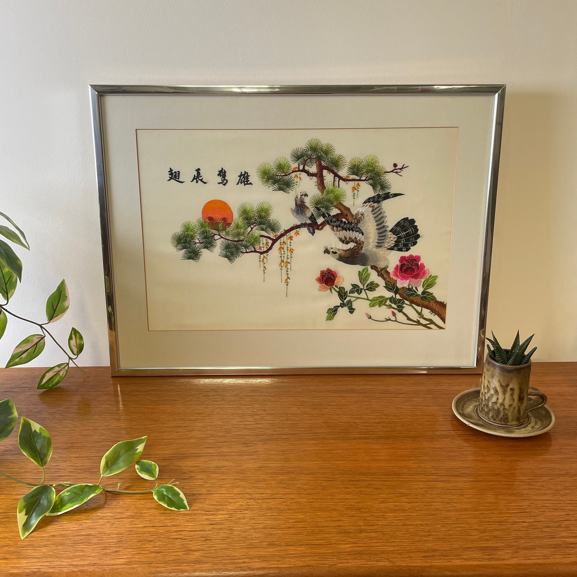 Vintage Silk Chinese Embroidered artwork in a silver and glass frame with a mounted card surround. On top of a teak unit with a ceramic potted succulent next to it. 