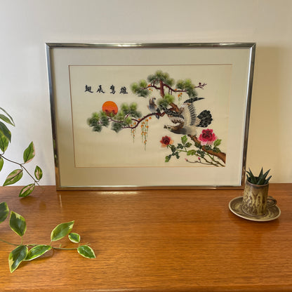 Vintage Silk Chinese Embroidered artwork in a silver and glass frame with a mounted card surround. On top of a teak unit with a ceramic potted succulent next to it. 