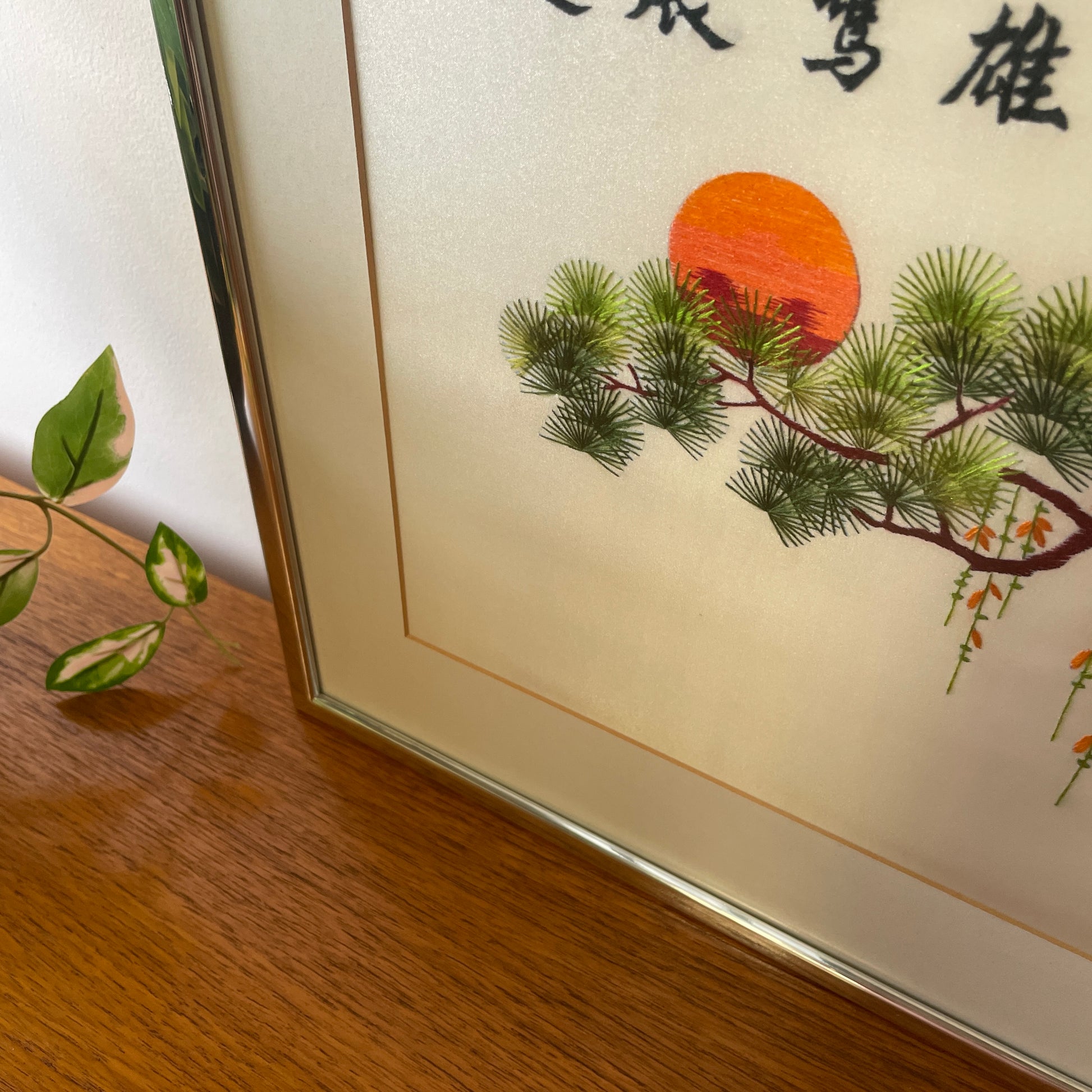 the silver frame, glass covering and card mount with a detailed shot of the embroidered art, green embroidered leaves against. red & orange embroidered sunset, with climbing trailing green & orange flowers falling from the tree. 