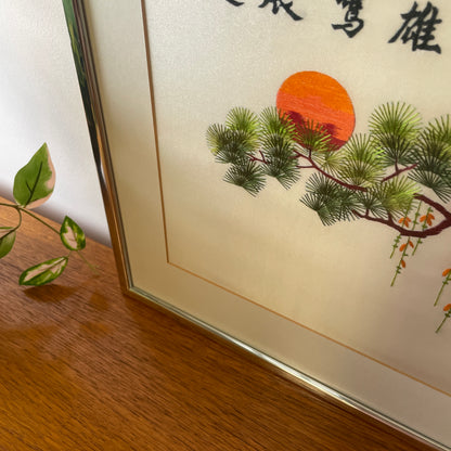 the silver frame, glass covering and card mount with a detailed shot of the embroidered art, green embroidered leaves against. red & orange embroidered sunset, with climbing trailing green & orange flowers falling from the tree. 