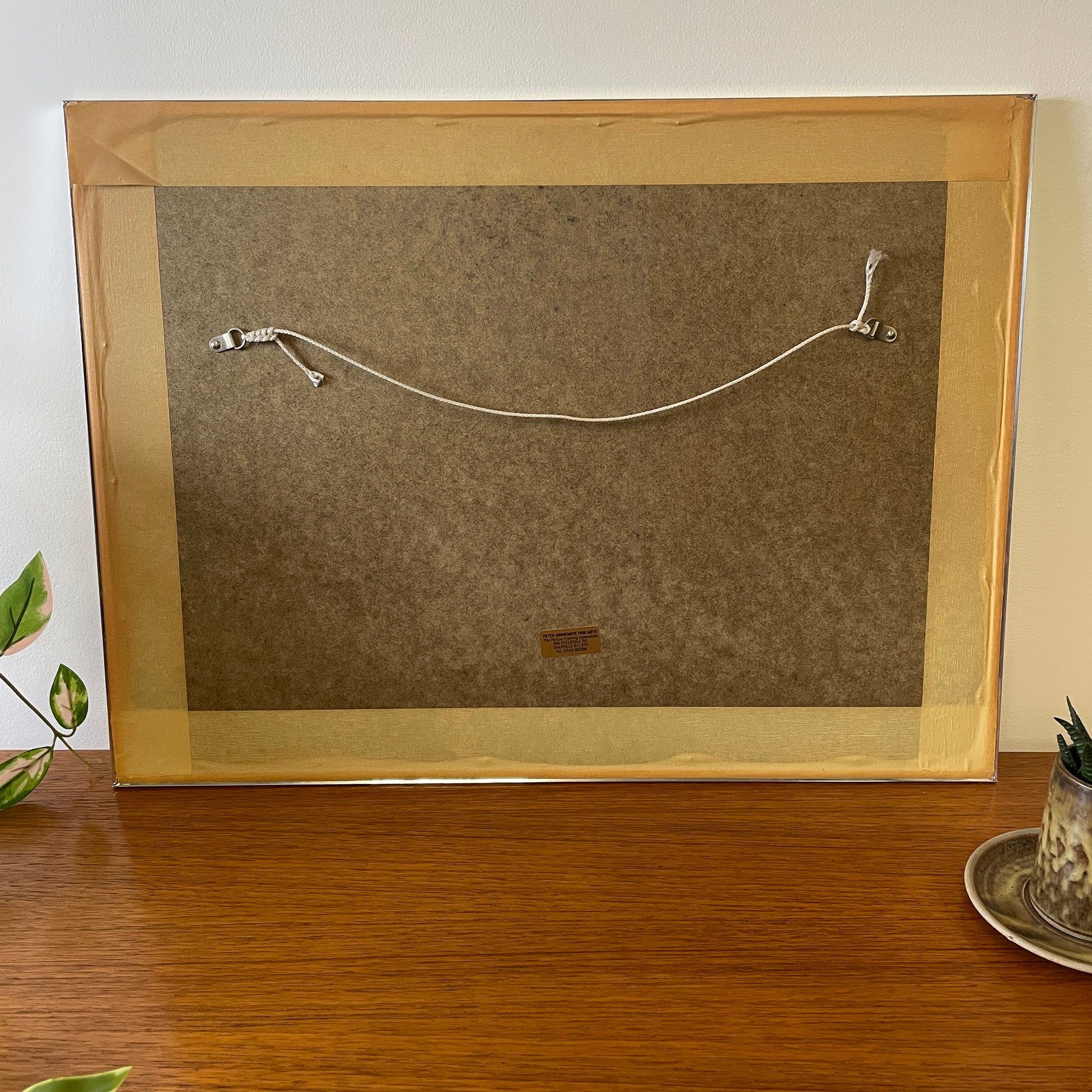 the back of the framed artwork is wood, with the original business name sticker of where it was made. It has string and metal clasps so it can be readily mounted and is secured in place with special tape to keep the artwork framed securely.