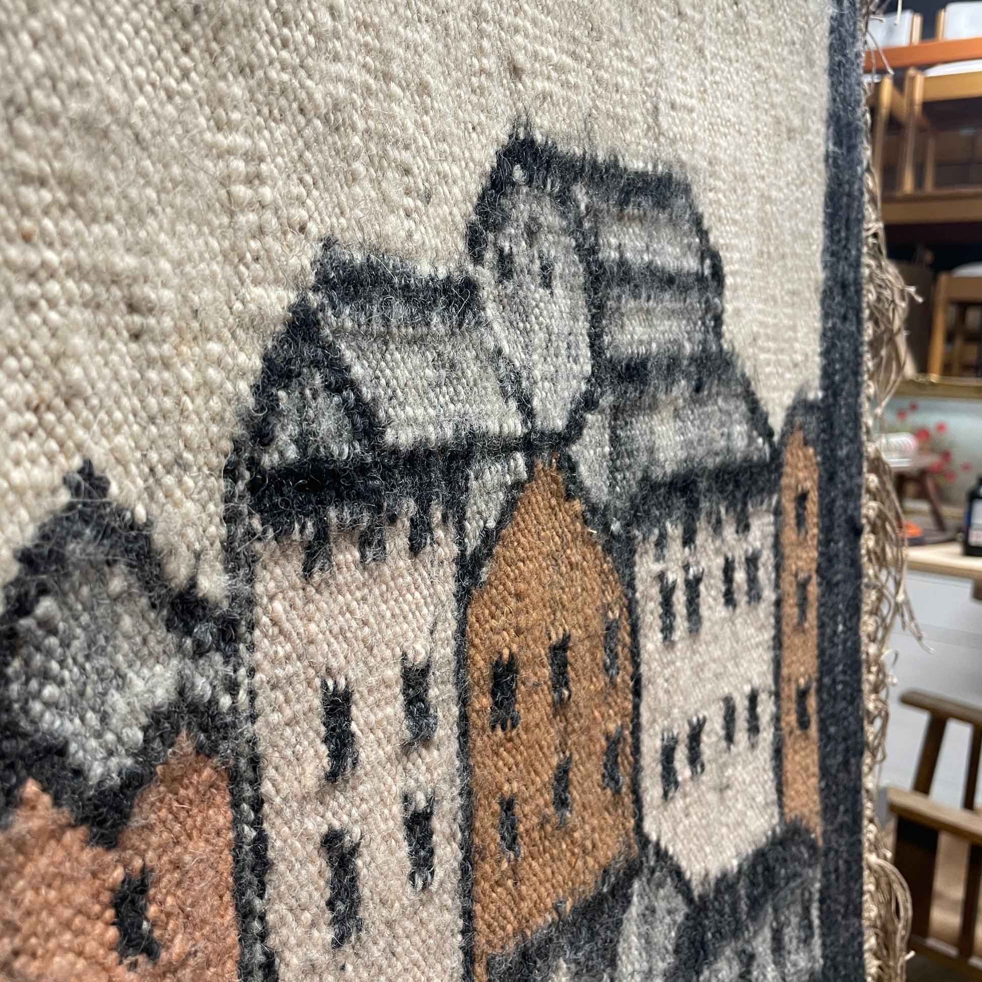 a closer up image of the weaved tapestry wall hanging workmanship, with the houses weaved in a clay black and cream colour against a cream background