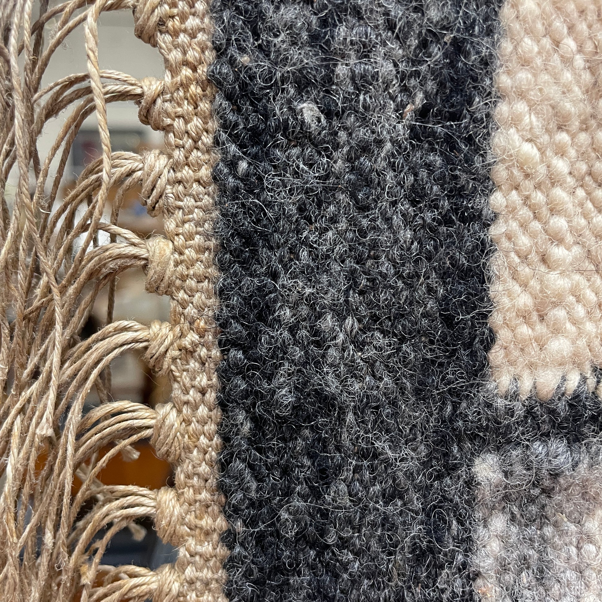 the side of the wall hanging tapestry, with a jute neutral colour surround and fringed edges knotted, and a black grey border shown. 